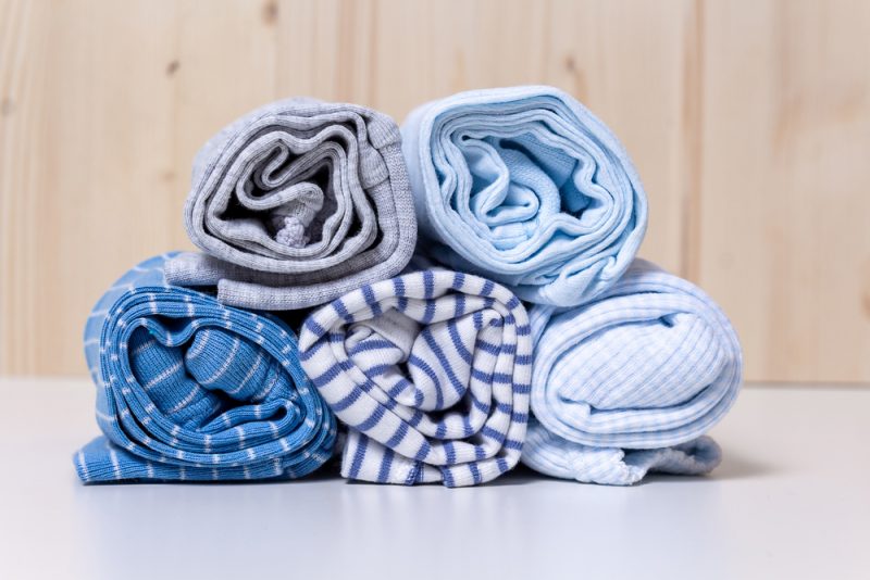 Clothes stack for newborn boy. Close up of rolled up kid's chlothes on white chest of drawers in front of wooden wall. Grey and blue colors and different print.